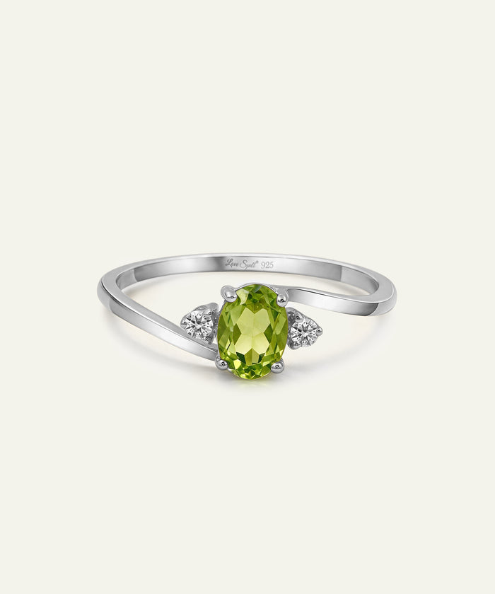 A silver ring featuring an oval-cut green peridot center stone and two small round-cut clear accent stones on either side of the band, shown on a light cream background.